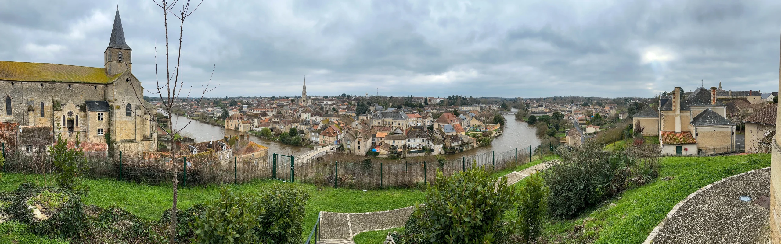 Panoramic stretch of French countryside with a medieval town on a river, under soft afternoon cloud cover — the slow shuttle between Slovakia and France where most of the real thinking gets done.