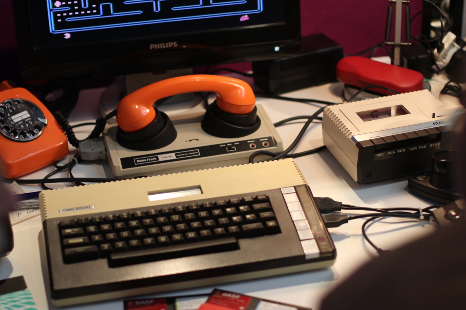 A vintage Atari 800XL home computer on a desk beside a 1980s acoustic-coupler modem with an orange telephone handset, an orange rotary dial phone and a cassette tape drive, a CRT monitor glowing faintly in the background.